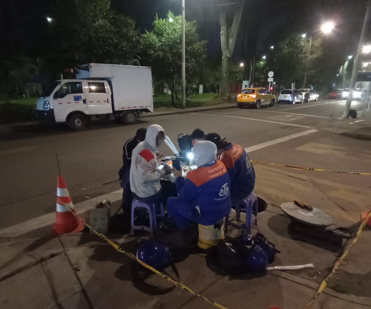 Un grupo de jóvenes técnicos de la ETB trabaja bajo la tenue luz de las farolas en una calle de Bogotá. Vestidos con chalecos reflectivos y cascos de seguridad, manipulan cables de fibra óptica, asegurando conexiones críticas mientras la ciudad duerme.